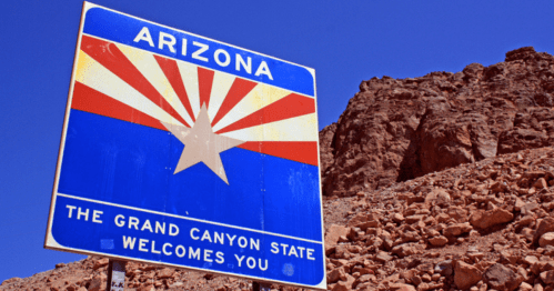 A welcome sign for Arizona featuring the state flag design, set against a rocky landscape under a clear blue sky.