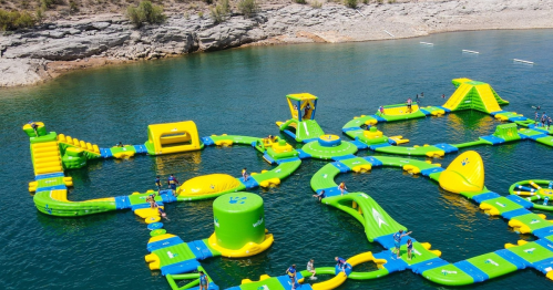 A colorful inflatable water park with slides and obstacles on a lake, surrounded by rocky shores and greenery.