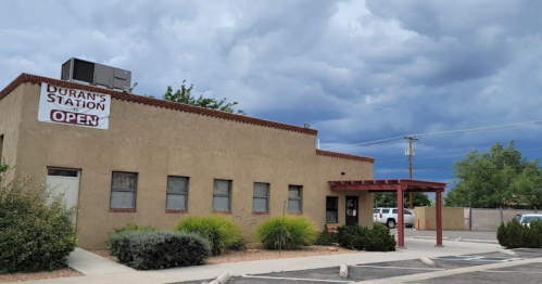 A small, tan building with a sign reading "Duran's Station OPEN" under a cloudy sky, surrounded by shrubs and a parking lot.