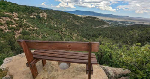 A wooden bench overlooks a lush valley and distant mountains under a partly cloudy sky.