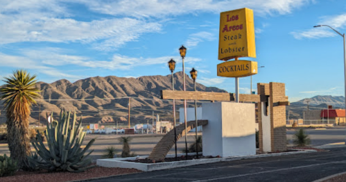 Sign for "Los Arcos Steak and Lobster" with mountains in the background and palm trees in the foreground.