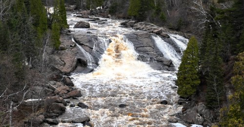 A rushing river cascades over rocky terrain, surrounded by trees and a hint of snow in a natural landscape.