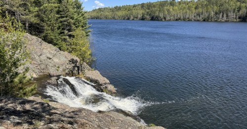 A serene river scene with a small waterfall cascading over rocks, surrounded by lush green trees and a clear blue sky.