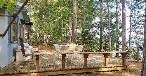 A wooden deck surrounded by trees, featuring a table and chairs, with a view of a lake in the background.