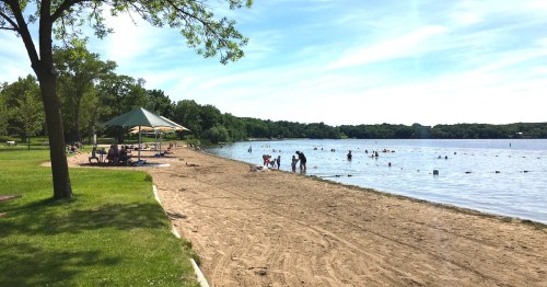 A sandy beach by a lake with people swimming, playing, and relaxing under a shaded area on a sunny day.