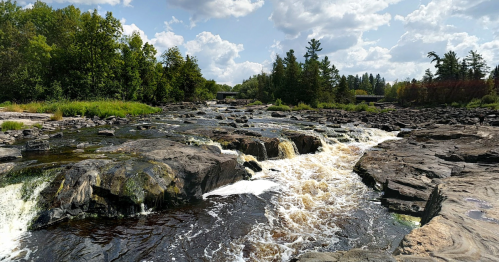 A serene river flows over rocky terrain, surrounded by lush greenery and a bright blue sky with scattered clouds.
