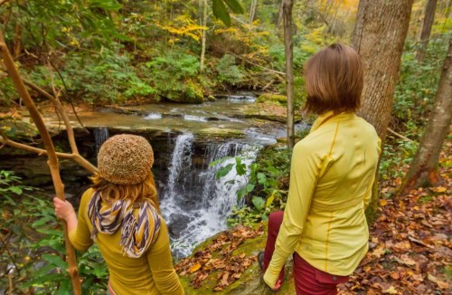 Two people in yellow jackets stand by a waterfall in a lush, autumn forest, surrounded by colorful foliage.