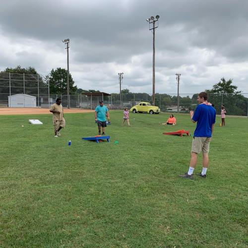 A group of people playing cornhole on a grassy field with a baseball diamond in the background under cloudy skies.