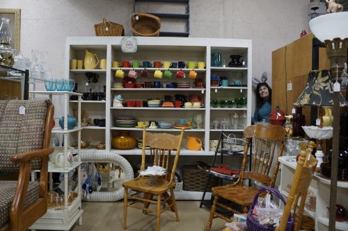 A vintage store display with colorful dishware and glassware, featuring wooden chairs and a woman with bunny ears in the background.