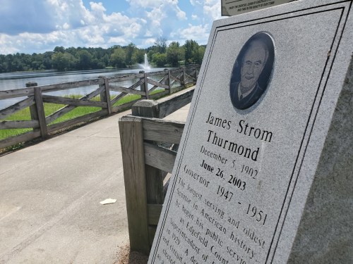 A stone plaque honoring James Strom Thurmond, with a lake and trees in the background on a sunny day.