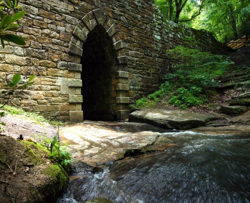 A stone archway bridge over a stream, surrounded by lush greenery and rocky terrain. Sunlight filters through the trees.