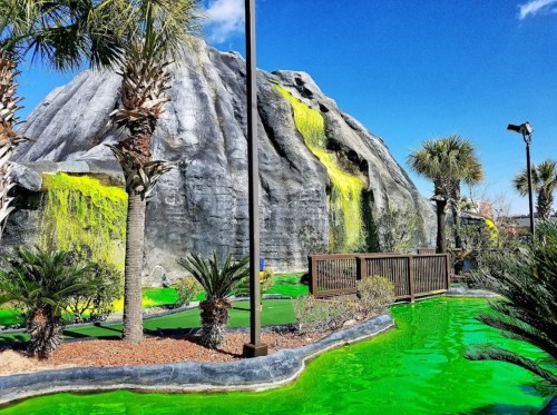 A mini-golf course featuring a large rock formation with waterfalls and vibrant green water surrounded by palm trees.