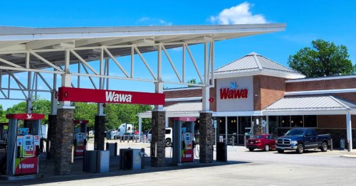 Red letters spelling Wawa above the entrance of a red brick convenience store with gas pumps under an outdoor canopy.