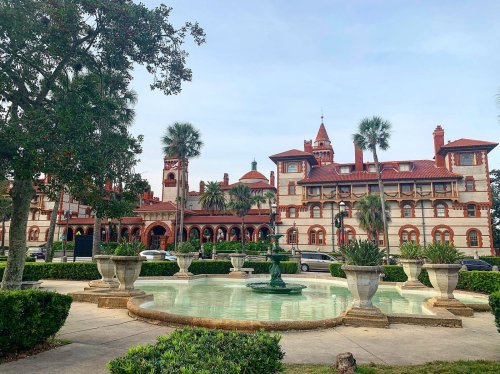 Historic building with red roofs and towers, surrounded by palm trees and a fountain in a landscaped area.