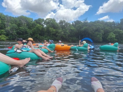 A group of people relaxing on colorful inflatable rafts in a river, surrounded by trees and a blue sky with clouds.