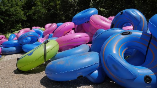 A colorful pile of inflatable tubes in blue, pink, and green, stacked on a gravel surface with greenery in the background.