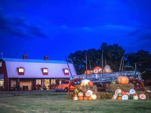A rustic farm scene at dusk, featuring a wagon with pumpkins and a red barn with lit windows in the background.