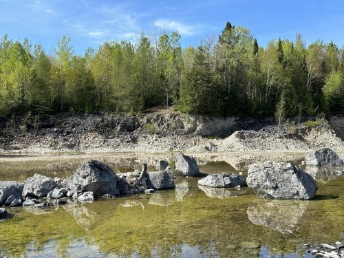 A serene landscape featuring a calm river with large rocks and lush green trees under a clear blue sky.