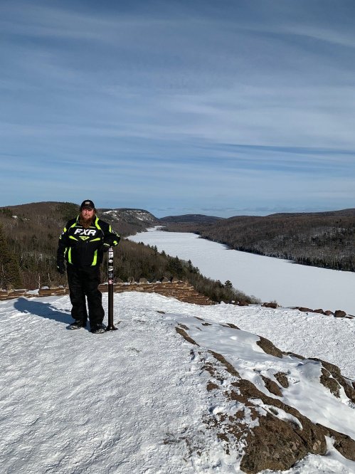 A person in winter gear stands on a snowy cliff overlooking a frozen lake and forested hills under a clear sky.