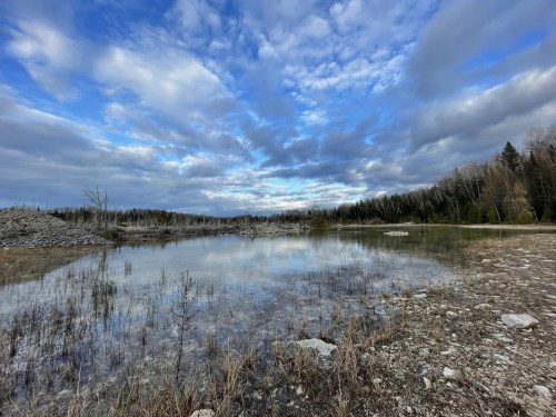 A serene landscape featuring a calm lake reflecting a cloudy sky, surrounded by trees and rocky terrain.