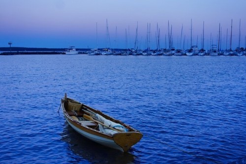 A small wooden boat floats in calm water, with a marina of sailboats in the background at dusk.