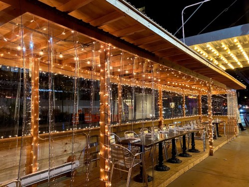 A cozy outdoor dining area with string lights and clear plastic barriers, creating a warm atmosphere at night.