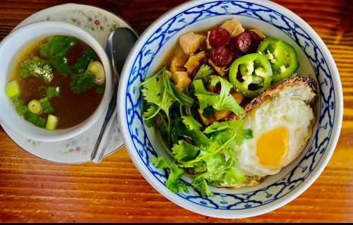 A bowl of rice topped with an egg, chicken, herbs, and jalapeños, alongside a cup of broth with green onions and cilantro.