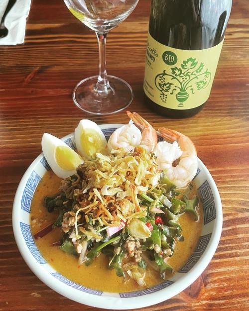 A bowl of vibrant soup with shrimp, boiled eggs, and crispy toppings, accompanied by a glass of white wine.