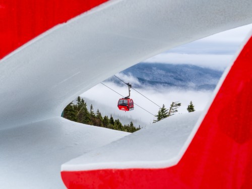 A red gondola travels above clouds, framed by a white and red sculpture in a mountainous landscape.