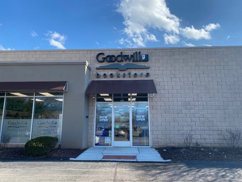 Exterior of a Goodwill bookstore with a "NOW OPEN" sign, featuring a tan brick facade and large glass entrance.