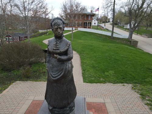 A statue of a woman in a dress stands in a park, with a historic building and trees in the background.