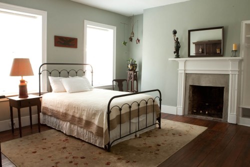 A cozy bedroom featuring a metal bed, a wooden chair, a fireplace, and soft natural light from large windows.
