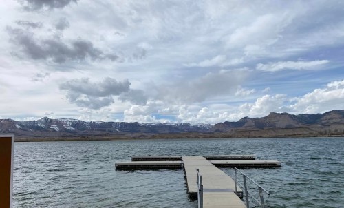 A wooden dock extends into a calm lake, surrounded by mountains and a cloudy sky.
