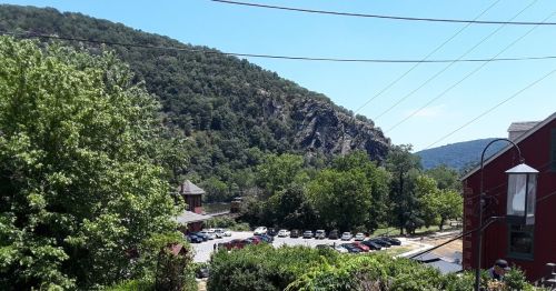 A scenic view of a green hillside with rocky cliffs, trees, and a parking area below under a clear blue sky.