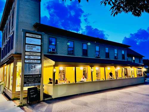 A cozy building at dusk with outdoor seating, featuring signs for local businesses and a welcoming atmosphere.