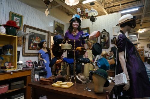 A vintage shop display featuring hats on stands, mannequins, and framed art in a charming, eclectic setting.