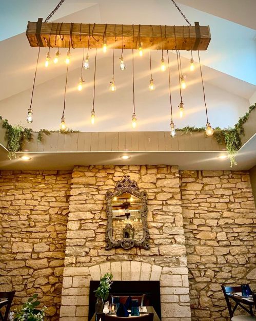 A rustic chandelier with hanging bulbs above a stone fireplace and a decorative mirror in a cozy dining space.