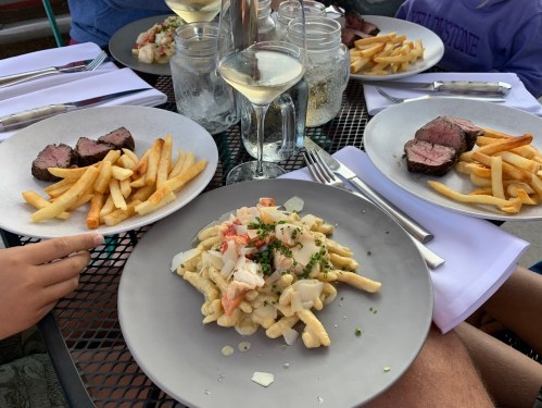 A table set with plates of steak, fries, and a seafood pasta dish, accompanied by glasses of white wine.