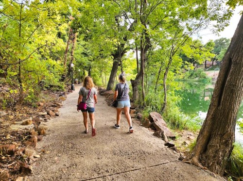 Two people walk along a tree-lined path by a calm lake, surrounded by greenery.