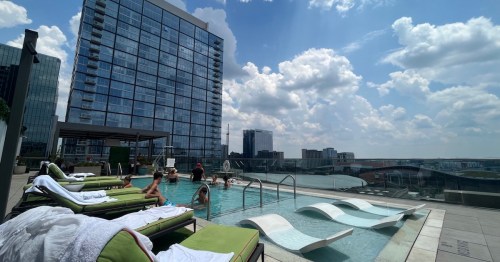 Rooftop pool scene with people relaxing, lounge chairs, and a city skyline under a partly cloudy sky.