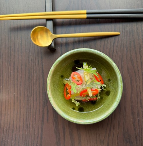 A green bowl with a colorful dish topped with sliced red chili and herbs, accompanied by gold utensils on a dark table.