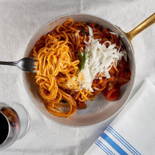 A serving of spaghetti in a silver pan, topped with cheese and accompanied by a glass of red wine on a white tablecloth.