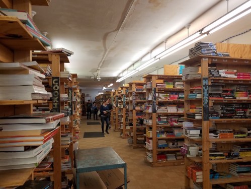 A cozy bookstore aisle lined with wooden shelves filled with various books, with people browsing in the background.