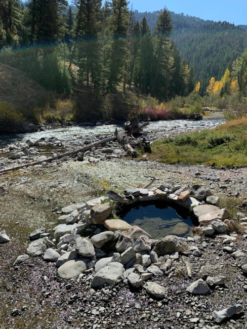 A natural hot spring surrounded by rocks, near a flowing river and forested mountains under a clear blue sky.