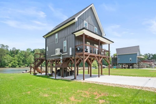 A modern two-story house on stilts, surrounded by green grass and trees, near a calm body of water.