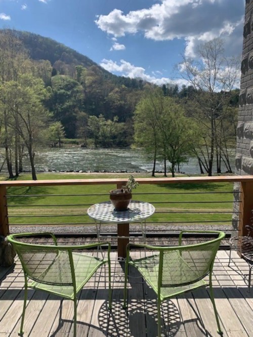 A scenic view from a deck featuring two green chairs and a small table overlooking a river and mountains.
