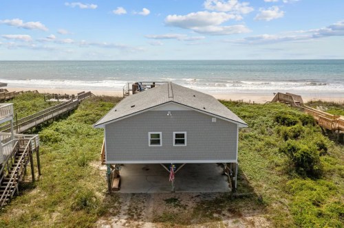 A gray beach house on stilts surrounded by greenery, with the ocean and blue sky in the background.