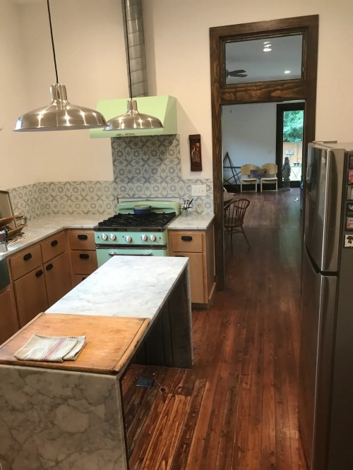 A modern kitchen with a green stove, marble island, and wooden floors, leading to a bright, open living space.