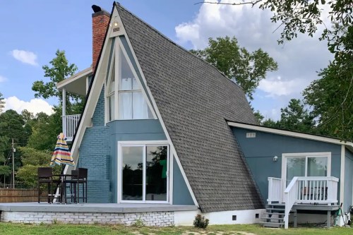A modern A-frame house with a sloped roof, large windows, and a small deck, surrounded by trees and greenery.