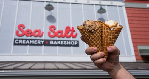 A hand holds three ice cream cones in front of the Sea Salt Creamery & Bakeshop sign.